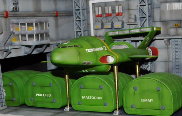 Thunderbird 2 elevated in hangar with pods underneath. Instead of the pods being numbered, they say "Pixelfed", "Mastodon", "Lemmy" on their doors. "Mastodon" is the pod currently underneath Thunderbird 2.
