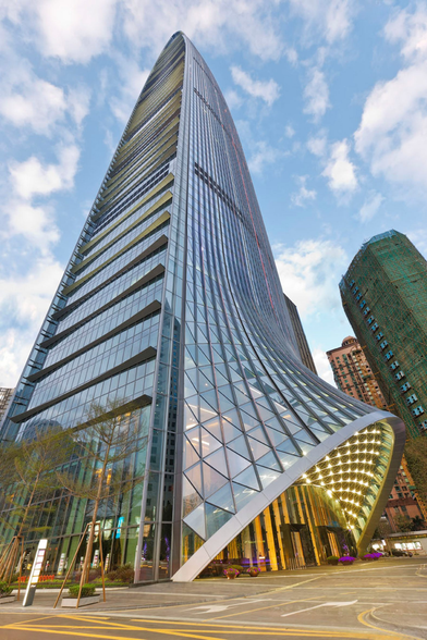 Modern high-rise skyscraper with a twisting glass and metal facade, curving dramatically at the base. The building features a geometric canopy with lights, surrounded by trees, other buildings, and a paved street with road markings.