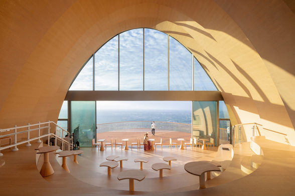 A contemporary interior space with a sweeping arched wooden ceiling and floor-to-ceiling windows overlooking the ocean. The room features tiered seating with sculptural wooden tables and chairs arranged in a semi-circle, while sunlight casts dramatic shadows across the curved surfaces. Outside, a wooden deck extends toward the water, where two people enjoy the coastal view.